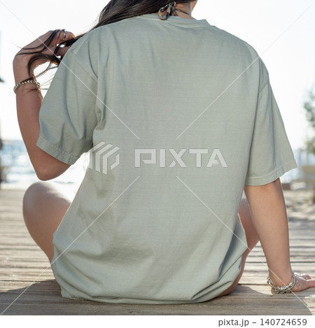 Sage green oversized t-shirt mockup, young woman sitting on beach boardwalk, back view, copy space Sage green oversized t-shirt mockup, young woman sitting on beach boardwalk, back view, copy space 140724659