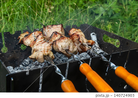 Grilling skewered meat over charcoal in a lush outdoor setting during a sunny afternoon Grilling skewered meat over charcoal in a lush outdoor setting during a sunny afternoon 140724903