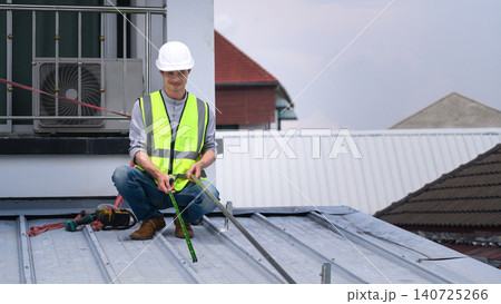 Asian Male Roofer in Safety Vest and Hard Hat Measuring Metal Roof During Construction Work Asian Male Roofer in Safety Vest and Hard Hat Measuring Metal Roof During Construction Work 140725266