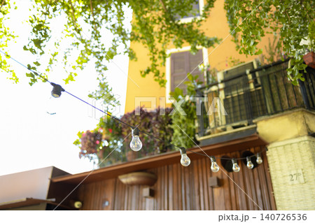 Cozy European building facade covered with green ivy and decorative outdoor string lights on a sunny day. Cozy European building facade covered with green ivy and decorative outdoor string lights on a sunny day. 140726536