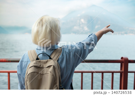 Joyful smiling mature elderly caucasian female traveler with backpack standing on promenade and pointing away towards scenic lake and mountains. Joyful smiling mature elderly caucasian female traveler with backpack standing on promenade and pointing away towards scenic lake and mountains. 140726538