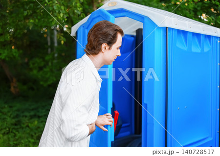 Man holding stomach in pain opening a blue outdoor portable toilet. Concept of an urgent toilet emergency, diarrhea, and digestive discomfort. Man holding stomach in pain opening a blue outdoor portable toilet. Concept of an urgent toilet emergency, diarrhea, and digestive discomfort. 140728517