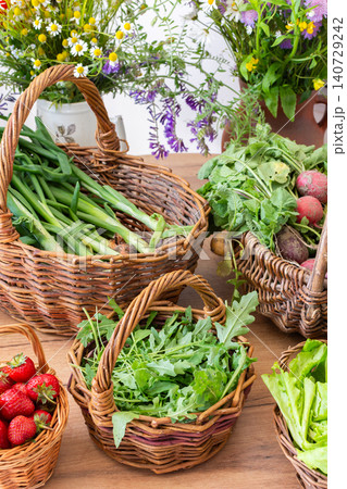 Freshly picked organic vegetables and strawberries are arranged in woven baskets on a wooden table. These local harvests show vibrant greens and red strawberries in a sunny setting 140729242
