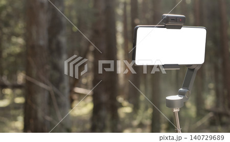 Smartphone mounted on a gimbal in a forest. Outdoor mobile filming setup with blurred trees in the background. Content creation, vlogging, and travel video concept.. Smartphone mounted on a gimbal in a forest. Outdoor mobile filming setup with blurred trees in the background. Content creation, vlogging, and travel video concept.. 140729689