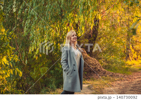Young blonde woman in grey coat walking park path enjoying golden autumn light Young blonde woman in grey coat walking park path enjoying golden autumn light 140730092