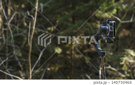 Smartphone mounted on a gimbal in a forest. Mobile filming gear stands against a woodland background. Outdoor content creation setup for vlogging and video recording.. Smartphone mounted on a gimbal in a forest. Mobile filming gear stands against a woodland background. Outdoor content creation setup for vlogging and video recording.. 140730468