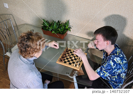 Two people playing chess at a table in a room with plants in the background during daytime hours Two people playing chess at a table in a room with plants in the background during daytime hours 140732107