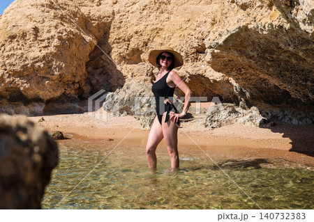 Young woman in a black swimsuit, hat, and sunglasses posing for the photographer against a stunning rocky seaside backdrop, capturing the essence of summer and beach vibes Young woman in a black swimsuit, hat, and sunglasses posing for the photographer against a stunning rocky seaside backdrop, capturing the essence of summer and beach vibes 140732383