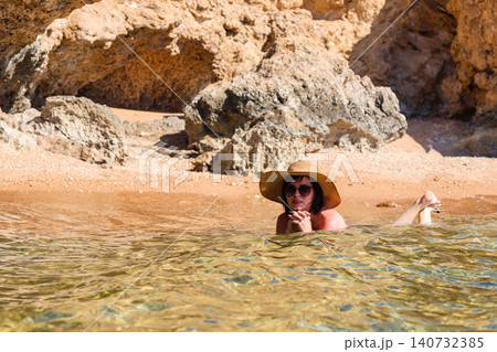Young woman in a black swimsuit, hat, and sunglasses posing for the photographer against a stunning rocky seaside backdrop, capturing the essence of summer and beach vibes Young woman in a black swimsuit, hat, and sunglasses posing for the photographer against a stunning rocky seaside backdrop, capturing the essence of summer and beach vibes 140732385