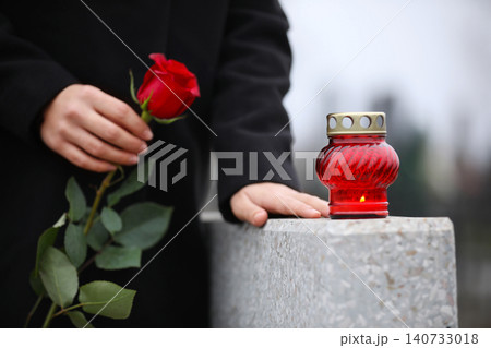 Woman holding red rose near grey granite tombstone with candle outdoors, closeup. Funeral ceremony Woman holding red rose near grey granite tombstone with candle outdoors, closeup. Funeral ceremony 140733018