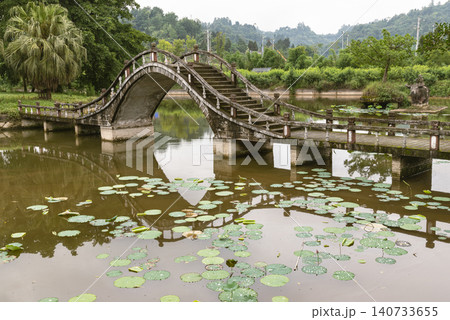 In Dazu Lotus Manor, a steeply stepped stone arch bridge vaults over a calm pond dotted with floating lotus foliage. Its high-arched silhouette casts a clear reflection onto the murky water, while a In Dazu Lotus Manor, a steeply stepped stone arch bridge vaults over a calm pond dotted with floating lotus foliage. Its high-arched silhouette casts a clear reflection onto the murky water, while a 140733655