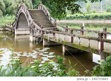 A timeless stone bridge arches over a tranquil, lily-dappled pond at Dazu Lotus Manor. The central span rises into a dramatic curve like a traditional Chinese moon bridge. Weathered steps climb A timeless stone bridge arches over a tranquil, lily-dappled pond at Dazu Lotus Manor. The central span rises into a dramatic curve like a traditional Chinese moon bridge. Weathered steps climb 140733665