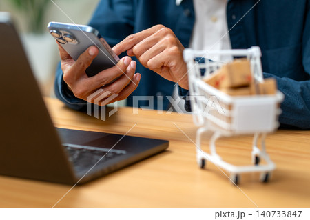 Close up hand of business man to hold mobile phone and use the other hand to touch on screen with cart containing small box is in front of business man. 140733847
