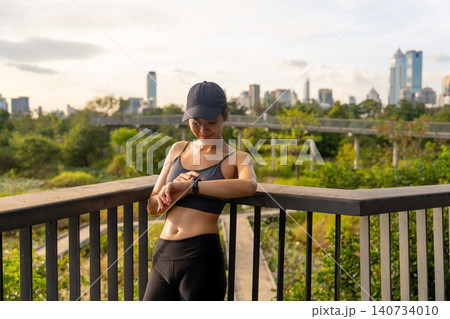 Asian sport woman is leaning on the railing and look on her smart watch with relax after workout in the big city central park. Asian sport woman is leaning on the railing and look on her smart watch with relax after workout in the big city central park. 140734010