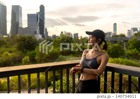 High view Asian sport woman is leaning on the railing and look on her right and smile with relax after workout in the big city central park. High view Asian sport woman is leaning on the railing and look on her right and smile with relax after workout in the big city central park. 140734011