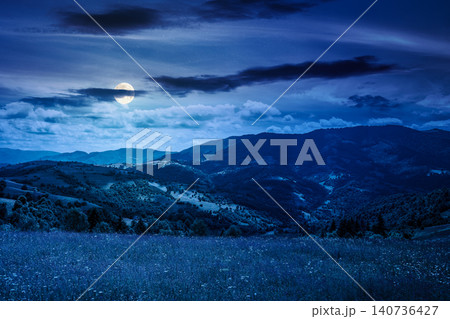 mountain landscape with grassy field at night. full moon over trees on the rolling hills and rural valley beneath a dark sky in the background. mysterious view of carpathian mountains in summer mountain landscape with grassy field at night. full moon over trees on the rolling hills and rural valley beneath a dark sky in the background. mysterious view of carpathian mountains in summer 140736427