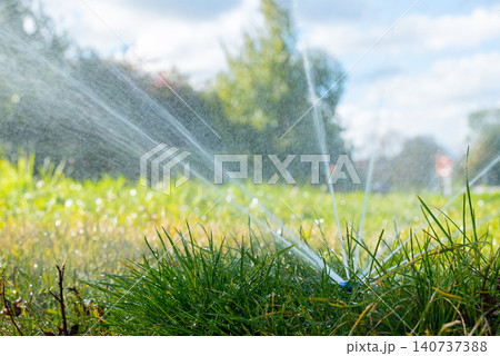 Automatic irrigation system sprays water across grassy lawn in outdoor garden environment. Fresh grass water droplets and irrigation hardware operating in residential or public green space. Natural 140737388