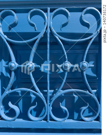 A close-up shot capturing a bold, bright blue painted metal grille with elegant, curved, and symmetrical scrollwork, set against a matching blue door to create a striking architectural detail in tradi 140738172
