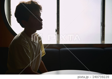 Soft backlight silhouettes a teenage boy at a table by a frosted window, suggesting solitude, reflection, travel and introspection in a calm, moody interior scene. Soft backlight silhouettes a teenage boy at a table by a frosted window, suggesting solitude, reflection, travel and introspection in a calm, moody interior scene. 140739220