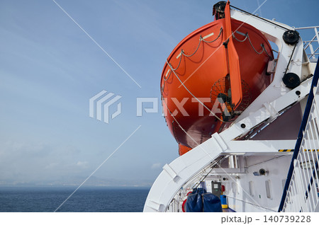 Bright orange lifeboat secured on a cruise-ship deck with davits and ropes, overlooking a calm blue sea under a clear sky; conveys travel, safety, maritime and rescue themes. 140739228
