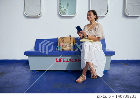 Smiling woman reads a passport while seated on a ferry bench next to lifejacket storage, enjoying a relaxed summer travel moment on deck with a straw bag and bright blue flooring. Smiling woman reads a passport while seated on a ferry bench next to lifejacket storage, enjoying a relaxed summer travel moment on deck with a straw bag and bright blue flooring. 140739233