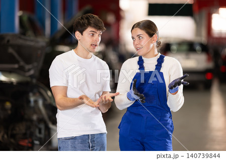 Male client talking to female mechanic in uniform about repairing his car engine in auto repair shop Male client talking to female mechanic in uniform about repairing his car engine in auto repair shop 140739844