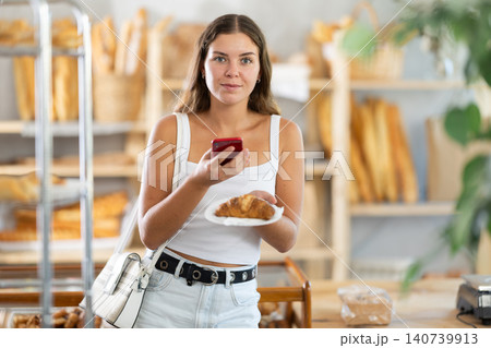 Young woman stands in bakery, scanning a croissant with her phone 140739913