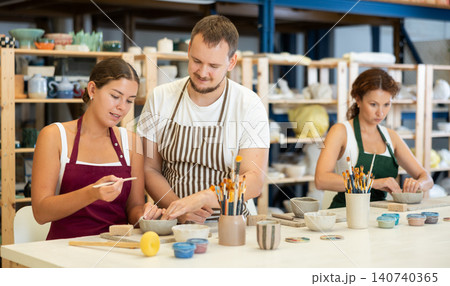 Male pottery teacher helps female students shape clay into plates and other dishes 140740365