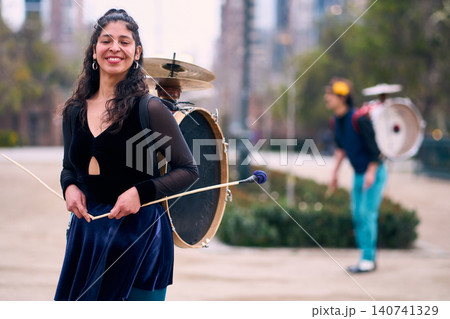 A beautiful, smiling Latina poses with her partner dancing in the background, carrying a drum and cymbals behind him. A Chinchinera, a street performer who dances and plays her drum in the streets A beautiful, smiling Latina poses with her partner dancing in the background, carrying a drum and cymbals behind him. A Chinchinera, a street performer who dances and plays her drum in the streets 140741329