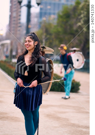 A beautiful, smiling Latina poses with her partner dancing in the background, carrying a drum and cymbals behind him. A Chinchinera, a street performer who dances and plays her drum in the streets A beautiful, smiling Latina poses with her partner dancing in the background, carrying a drum and cymbals behind him. A Chinchinera, a street performer who dances and plays her drum in the streets 140741330