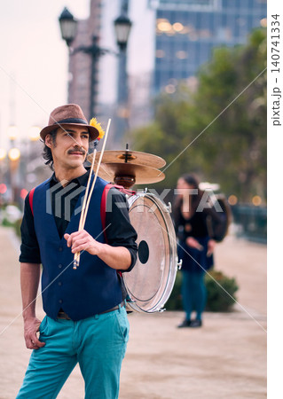 Adult man in colorful clothing poses cheerfully and stylishly, with his dancing partner in the background. A Chilean street musician, known as a chinchinero, plays the bass drum and dances Adult man in colorful clothing poses cheerfully and stylishly, with his dancing partner in the background. A Chilean street musician, known as a chinchinero, plays the bass drum and dances 140741334