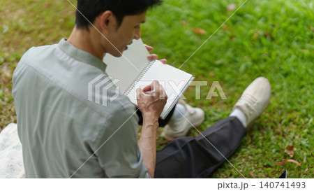 Asian man writing in notebook with pen in park for journaling 140741493