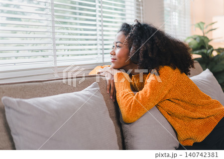 Thoughtful African American woman, resting on a couch, gazing out of a window with a serene expression. Thoughtful African American woman, resting on a couch, gazing out of a window with a serene expression. 140742381