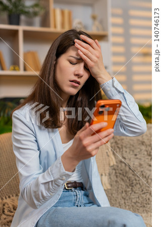 Young woman on home sofa reading bad news on smartphone showing shock stress upset reaction indoors 140746173