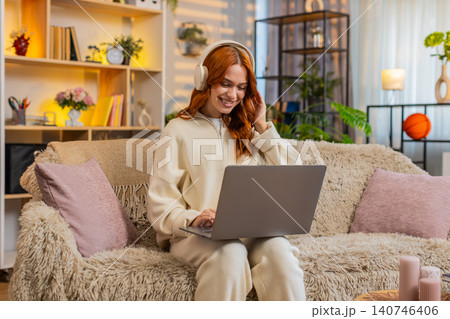 Young woman on home sofa working on laptop with headphones smiling to music during home office work 140746406