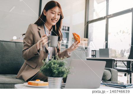 Young asian businesswoman eating a brownie while working from her office on a laptop next to a window with blinds Young asian businesswoman eating a brownie while working from her office on a laptop next to a window with blinds 140746639