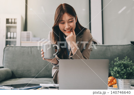 Young asian businesswoman eating a brownie while working from her office on a laptop next to a window with blinds 140746640
