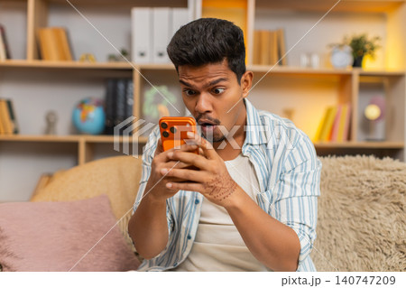 Indian man on home sofa reading bad news on smartphone and covering face in despair in living room Indian man on home sofa reading bad news on smartphone and covering face in despair in living room 140747209
