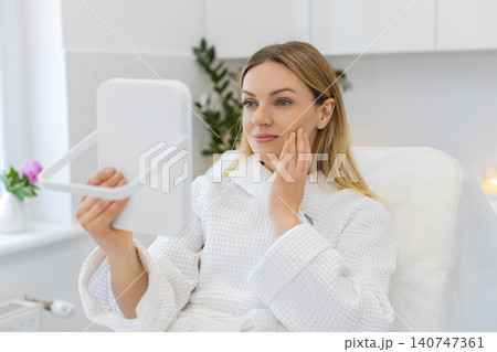 Caucasian young woman smiles in mirror after facial therapy in beauty clinic showing visible result Caucasian young woman smiles in mirror after facial therapy in beauty clinic showing visible result 140747361