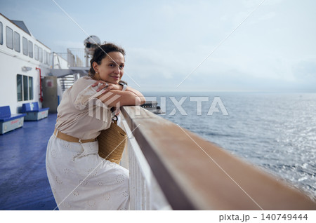 A smiling woman leans on a ferry railing, gazing across the calm sea with a straw bag and light dress. Soft sunlight and open horizon evoke relaxed travel, leisure and seaside reflection. A smiling woman leans on a ferry railing, gazing across the calm sea with a straw bag and light dress. Soft sunlight and open horizon evoke relaxed travel, leisure and seaside reflection. 140749444
