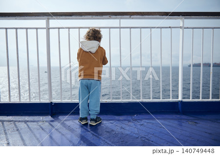 Young boy stands at a ferry railing, gazing across calm open sea toward the distant horizon. The scene suggests travel, solitude, childhood curiosity and a peaceful coastal journey on a bright day. Young boy stands at a ferry railing, gazing across calm open sea toward the distant horizon. The scene suggests travel, solitude, childhood curiosity and a peaceful coastal journey on a bright day. 140749448
