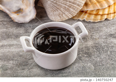 White ceramic bowl filled with dark sauce beside seashells on textured surface 140750247