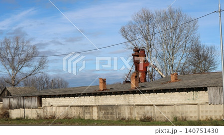An abandoned Soviet-era barn made of sand-lime blocks with a slate roof and crumbling brick chimneys. A rusty cyclone grain bin towers behind a concrete fence. Bare trees and dead wood surround it. An abandoned Soviet-era barn made of sand-lime blocks with a slate roof and crumbling brick chimneys. A rusty cyclone grain bin towers behind a concrete fence. Bare trees and dead wood surround it. 140751430