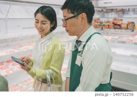 スーパーの精肉売り場でスマホを見せて満面の笑みで話す女性客と無愛想な店員 140752256