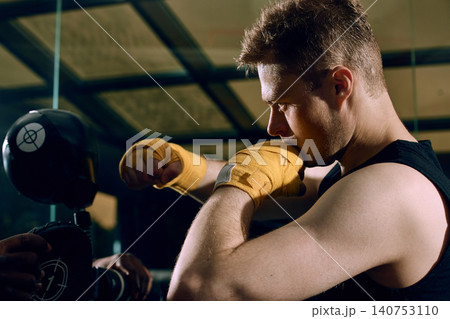 Focused boxer with yellow hand wraps punching reflex bag in dark gym. Focused boxer with yellow hand wraps punching reflex bag in dark gym. 140753110