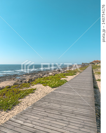 A beautiful long wooden boardwalk stretches alongside the rocky Atlantic shore and sandy dunes of Praia da Granja in Porto Portugal under a bright clear blue sky during a sunny summer day A beautiful long wooden boardwalk stretches alongside the rocky Atlantic shore and sandy dunes of Praia da Granja in Porto Portugal under a bright clear blue sky during a sunny summer day 140754176