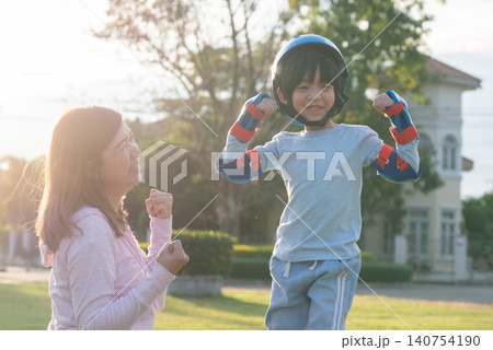 Happy Asian mother encouraging her little son while he is putting on a safety helmet and protective gear at the park. Happy Asian mother encouraging her little son while he is putting on a safety helmet and protective gear at the park. 140754190
