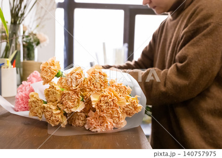 Woman florist wrapping bouquet of peach carnation flowers. Professional florist working in flower shop. Floral designer preparing fresh carnation arrangement for celebration and gift. Woman florist wrapping bouquet of peach carnation flowers. Professional florist working in flower shop. Floral designer preparing fresh carnation arrangement for celebration and gift. 140757975