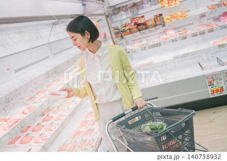 スーパーの精肉売り場でパック肉を手に取り品定めする女性客と野菜入り買い物かご 140758932