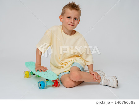 A handsome young boy sitting on a white studio floor, leaning his arm on a mint green penny board. Minimalist commercial photography showcasing children's summer hobbies and casual street fashion A handsome young boy sitting on a white studio floor, leaning his arm on a mint green penny board. Minimalist commercial photography showcasing children's summer hobbies and casual street fashion 140760388
